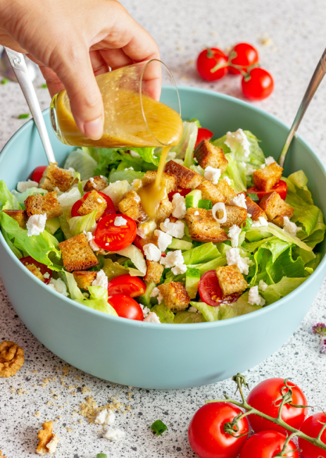A healthy salad is kept on a marble surface and a person is pouring salad dressing into the bowl of salad.